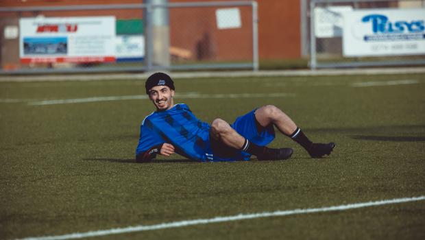 Football player in black beanie cap and blue kit lying on the grass smiling