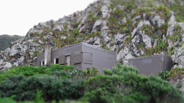 Grey shack on beach at higher ground surrounded by mountains and foliage