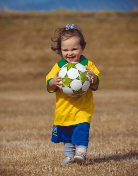 Girl in Brasil kit and white black dotted hair bow carrying a football - Little Dribblers