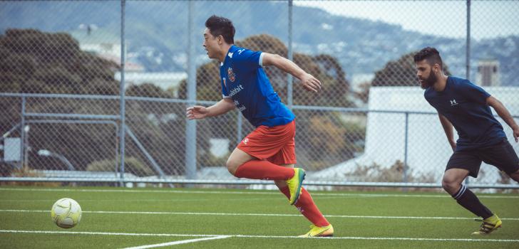Asian player in orange shorts chasing after football - Sports Zone sunday league