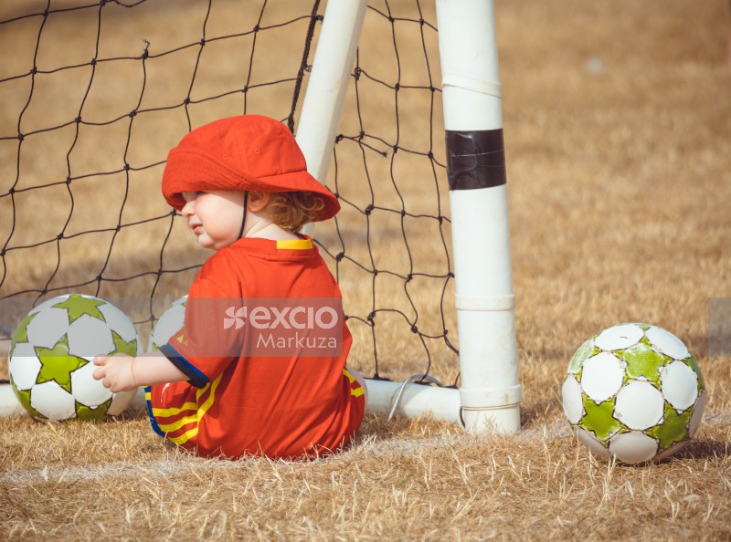 Little girl wearing a red hat sitting inside a goal post - Little Dribblers
