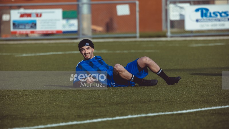 Football player in black beanie cap and blue kit lying on the grass smiling