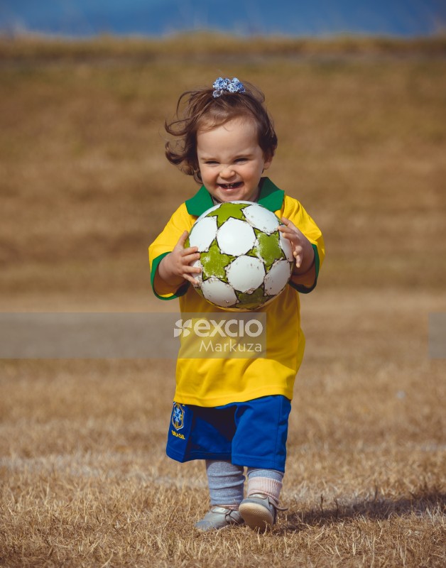Girl in Brasil kit and white black dotted hair bow carrying a football - Little Dribblers