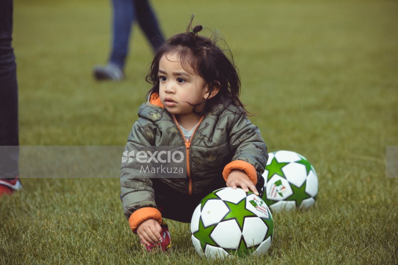 Girl in a dinosaur drawn jacket sitting in the grass with footballs - Little Dribblers