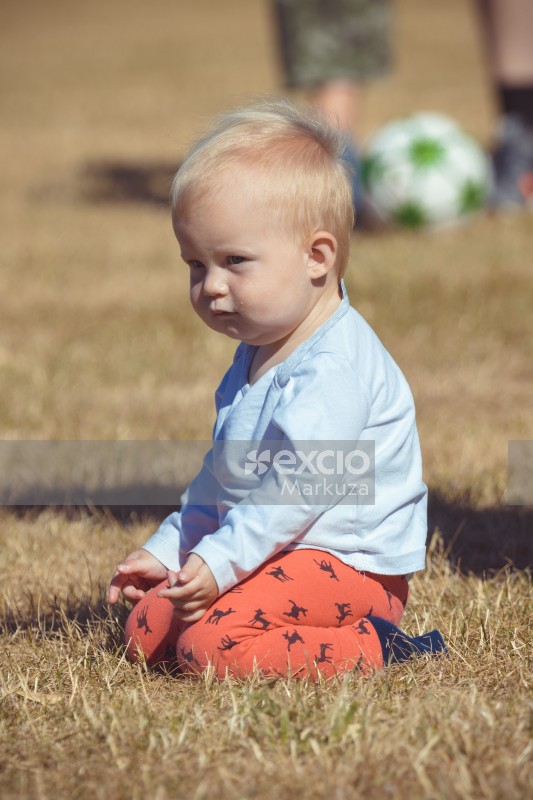 Blonde toddler sitting in dry grass at Little Dribblers football game