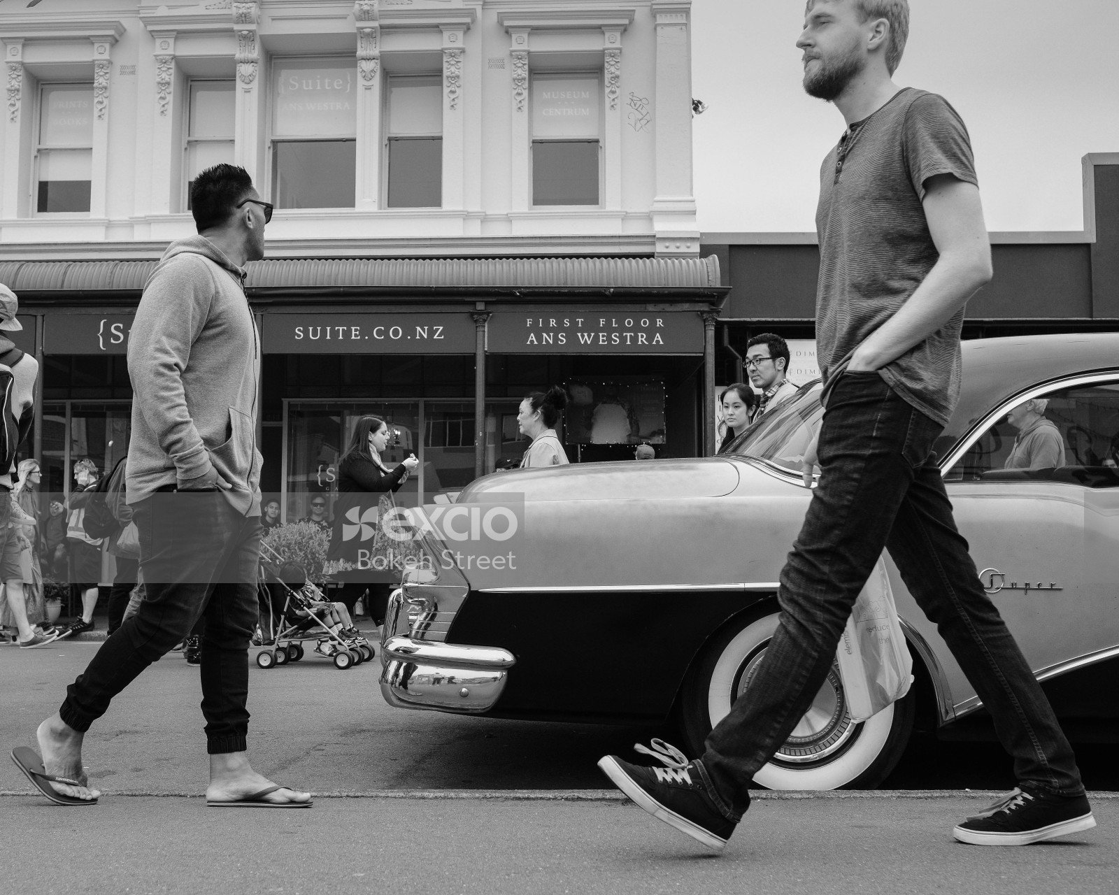 People walking by a classic car parked in the street at a festival black and white