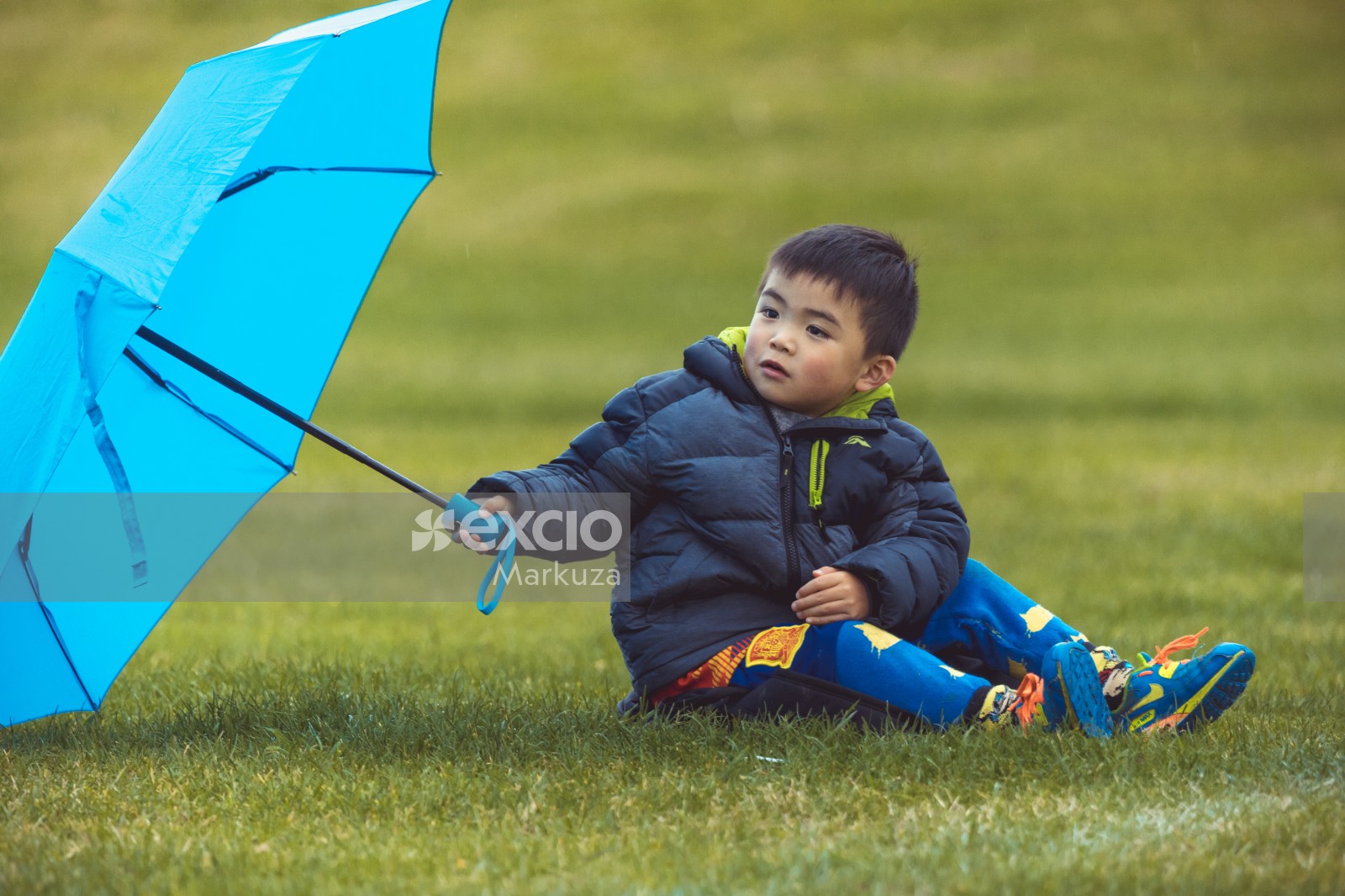 Boy with a blue umbrella sitting on grass at Little Dribblers soccer contest