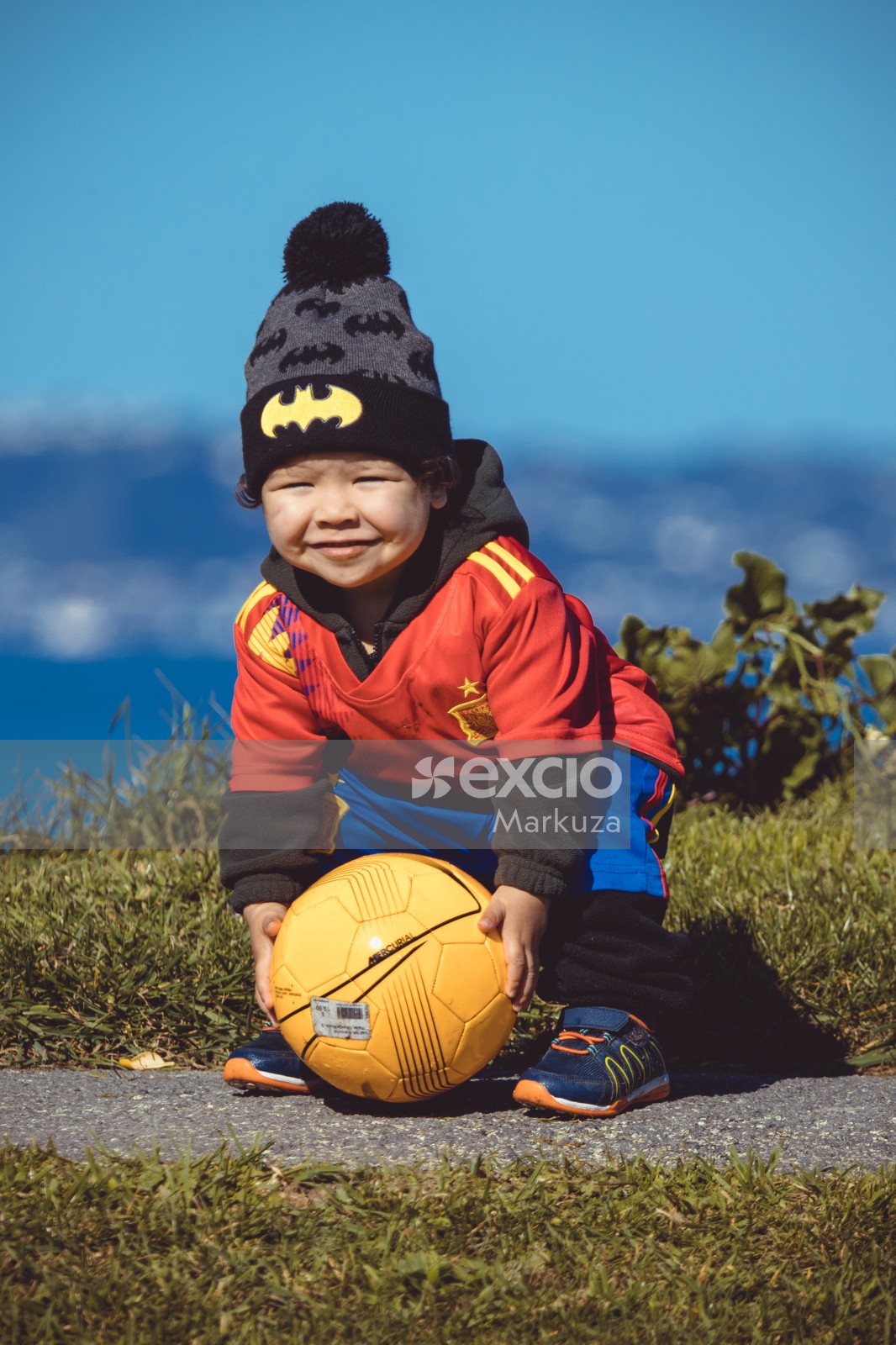 Kid in Manchester United kit and batman cap holding a yellow football - Little Dribblers