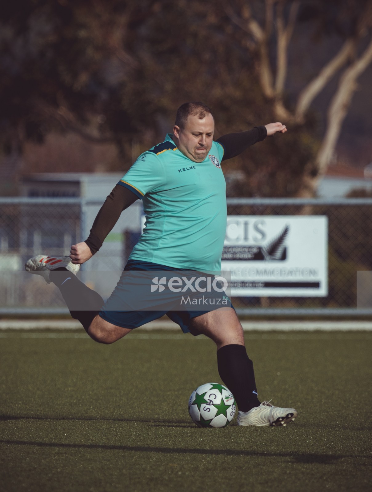 Chubby football player in turquoise Kelme shirt kicking ball